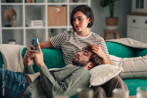 Bild auf Leinwand Young couple relaxing at home on a couch, man using smartphone while woman cares