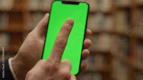 Close-up of a persons hands holding and interacting with a smartphone featuring a vibrant green screen, set against a blurred background of bookshelves, ideal for technology and education themes.