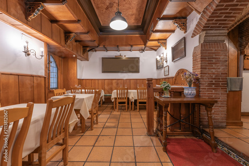 Traditional wood-beamed dining room with white-clothed tables, a brick archway, and decorative pottery near a red rug entrance.