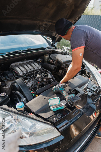 Mechanic inspecting car engine under open hood at outdoor workshop, using diagnostic tools with focused posture during routine maintenance and repair work, with mechanic, inspecting, engine, hood