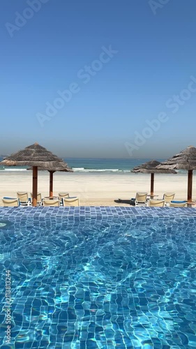Infinity swimming pool with sun loungers and straw umbrellas facing pristine beach and clear blue sea. Elegant coastal resort creates peaceful holiday atmosphere