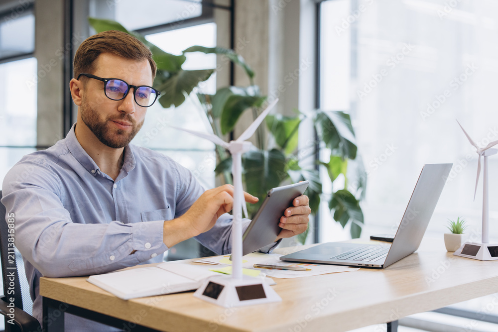 Obraz premium Male engineer using a tablet, working on renewable energy project with wind turbine models in a modern office