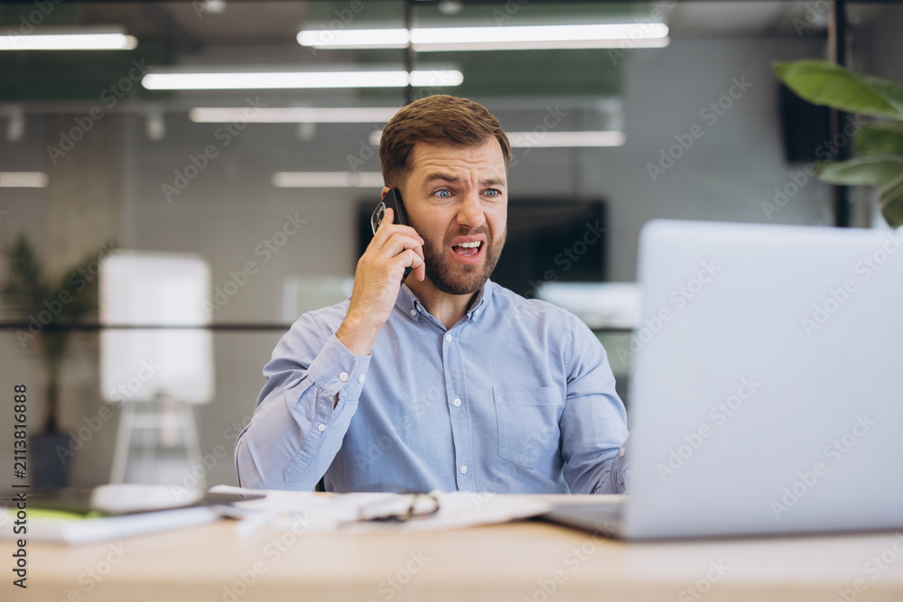 Fototapeta premium Stressed businessman talking on smartphone, reacting with anger and frustration to bad news, working on laptop in modern office