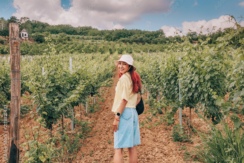 Obraz premium Smiling woman exploring lush green vineyard rows on a sunny summer day. Travel concept.