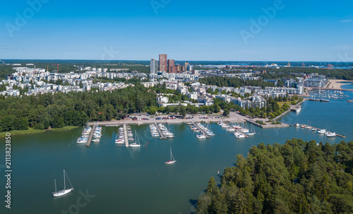 Elevated panoramic view of Vuosaari district in Helsinki, Finland, featuring a sailboat marina, modern apartment buildings, pine forest coastline, and high-rise towers on the horizon