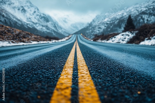Low angle perspective of a wet asphalt highway leading toward snowy mountain peaks under a cloudy sky