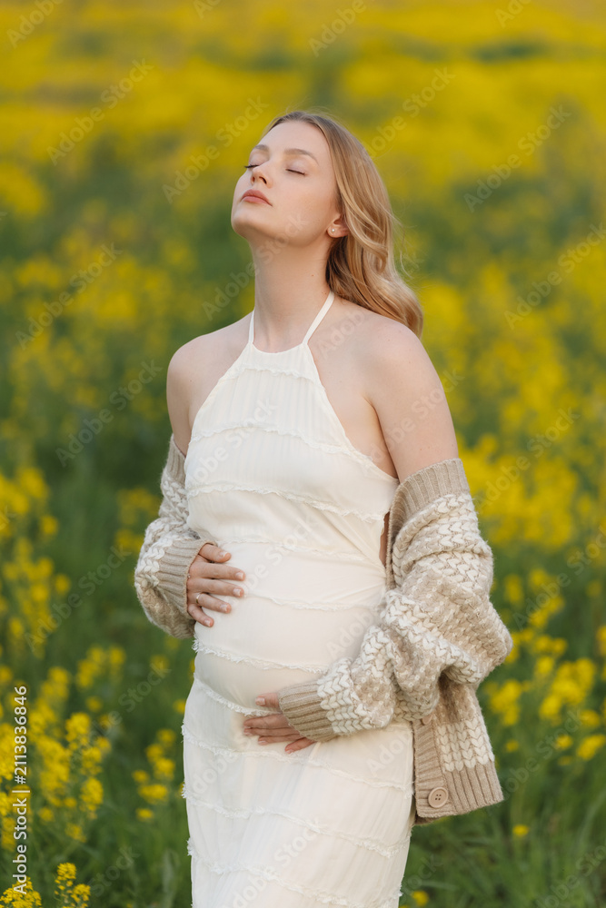 Obraz premium Pregnant woman in flowing white dress and knitted cardigan stands in yellow flower meadow with eyes closed and hands on belly, showing calm anticipation, gentle natural light, blurred spring