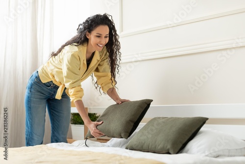 Quadro su tela Smiling Black Woman In Her 30s Arranging Pillows On Bed In Cozy Bedroom, Bringing Comfort And Style To Her Living Space, Making It Inviting