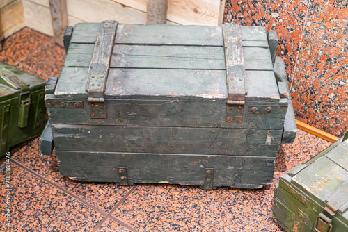 Weathered military-style wooden box with dark green paint, positioned on a polished terrazzo floor, showing metal latches and hinges, slightly rusted, surrounded by wooden wall elements, indoor