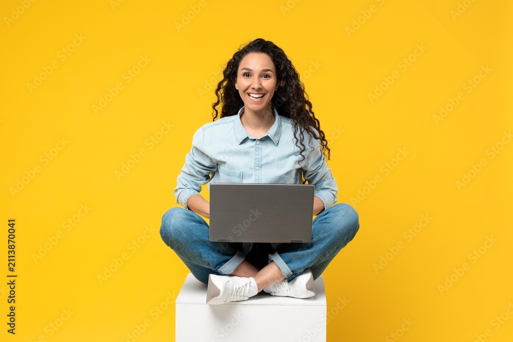 Fototapeta premium Smiling Young Mixed Race Woman Sitting Cross-legged On Cube While Working On Laptop In Studio With Bright Yellow Background. Joyful Emotion Conveyed In A Modern Remote Work Concept.
