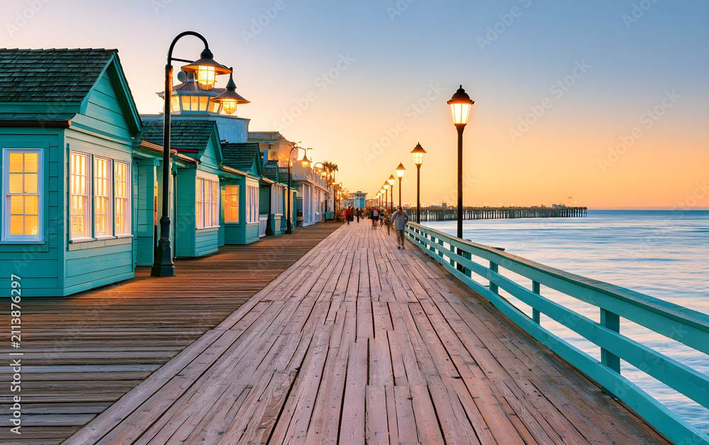 Fototapeta premium Oceanfront boardwalk with light blue buildings, shops and lamp posts at sunset. People walk along the wooden pier near the sea. Relaxing coastal vacation scene