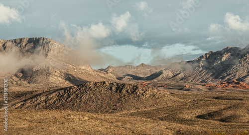Obraz na plátně Fog and haze soften desert mountains under an overcast sky, with reddish rock outcrops visible along the base of the slopes