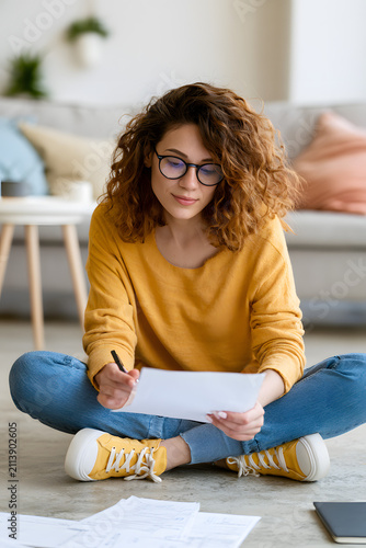 A vertical image of a young adult Caucasian woman seated on the floor, assessing her bills and notes as she manages household finances and copes with financial stress. Perfect for promoting informatio