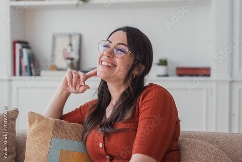 Foto A happy young woman lounges on a sofa in a bright living room, smiling and looking thoughtful while wearing glasses and a rust-colored sweater, conveying comfort, relaxation, and contentment