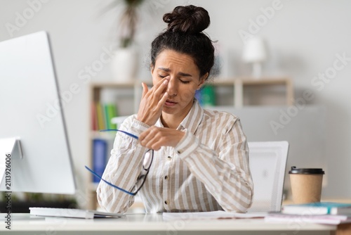 Bild auf Leinwand Worried Hispanic Female Professional Rubbing Her Eyes While Working At Desk In Modern Office Setting