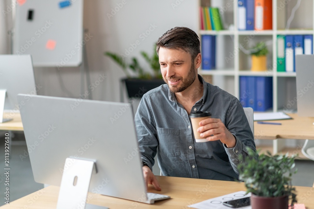 Obraz premium Smiling Caucasian Man In His 30s Working On Computer While Enjoying Coffee In Bright Modern Office Environment. Focused On Task, Creating Content, And Productive Atmosphere In Workspace.