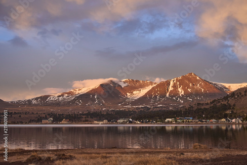 Winter landscape of Caviahue with snow covered Andes mountains at sunrise, in Neuquen, Argentina. The houses in the tourist town are reflected in the lake in the foreground.