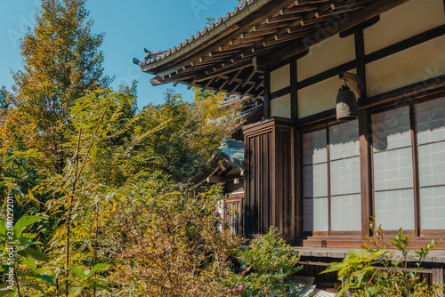 Traditional Japanese House in Autumn: An ancient Japanese wooden house with characteristic tiled roof and shoji screens stands amidst lush greenery and colorful autumn foliage.