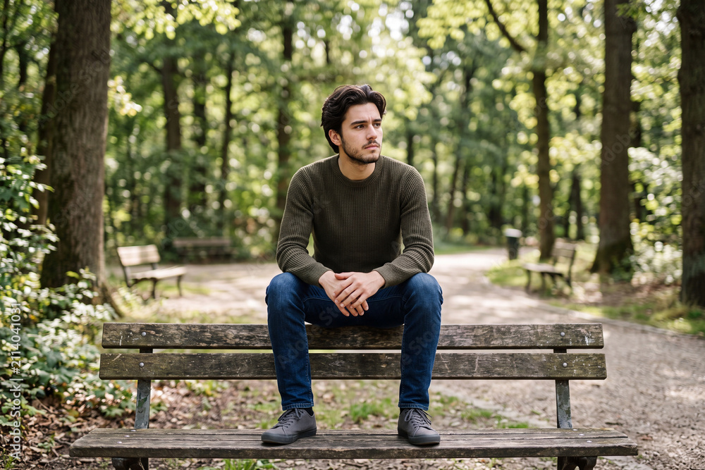 Fototapeta premium Young Man Sits Alone on Park Bench Lost in Deep Thought and Quiet Internal Struggle