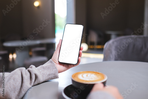 Mockup image of a woman holding mobile phone with blank desktop screen while drinking coffee in cafe