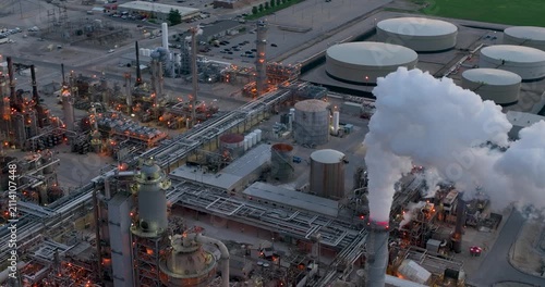 Aerial view of an oil refinery at sunset with storage tanks, industrial infrastructure, and smoke rising 