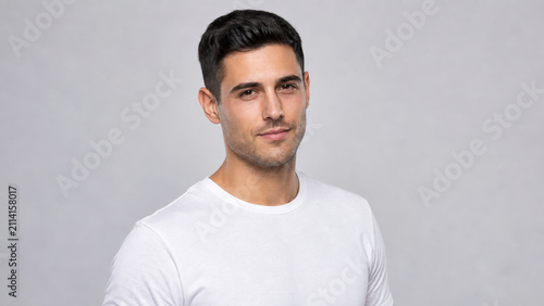 Young man with short dark hair wearing white t shirt posing confidently with slight smile on gray background