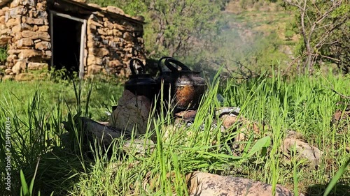 A rustic stone hut nestles into a green hillside near towering rocky mountains, while kettles simmer over a smoky campfire in the grass below.
📍 Hawraman Takht, Kurdistan, Iran 