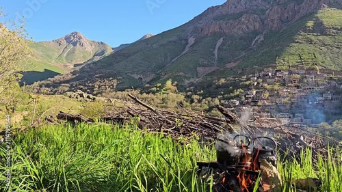 Blackened kettles steam over a crackling open campfire amid tall green grass, with a sunlit rocky mountain and distant village unfolding beyond.
📍 Kamalla village, Hawraman Takht, Kurdistan 