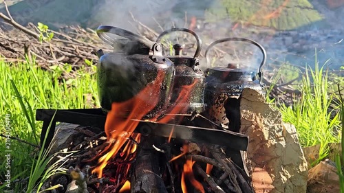 Blackened kettles steam over a crackling open campfire amid tall green grass, with a sunlit rocky mountain and distant village unfolding beyond.
📍 Kamalla village, Hawraman Takht, Kurdistan 