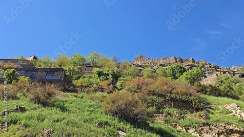 Traditional stone houses perch on a green hillside beneath jagged rock formations, surrounded by wild shrubs under a clear, expansive blue sky.
📍 Hawraman Takht, Kurdistan, Iran 