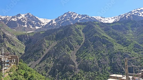 Rugged snow-capped peaks tower over a terraced stone village and forested valley, with a mountain road winding through the greenery below a clear blue sky.
📍 Hawraman Takht, Kurdistan, Iran 