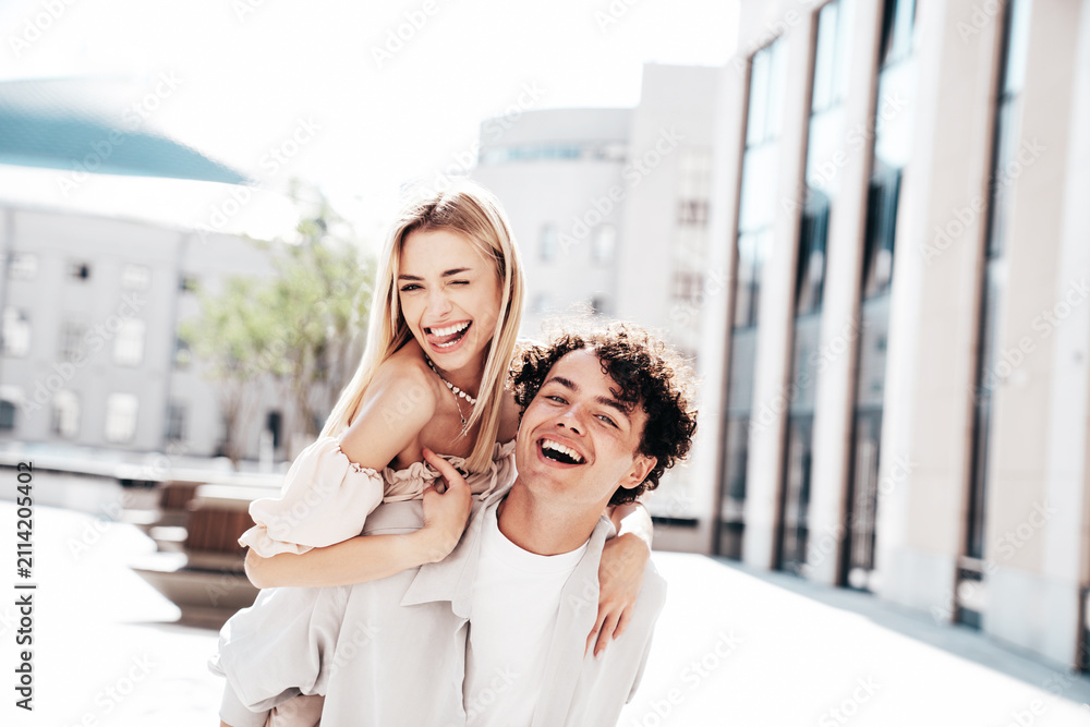 Obraz premium Young smiling beautiful woman and her handsome boyfriend in casual summer clothes. Happy cheerful family. Couple posing in street at sunny day. Female sits at his back. piggyback riding