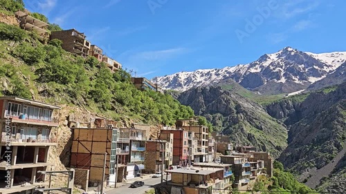 On a steep mountainside, a picturesque village of stacked stone houses overlooks a winding road, lush green slopes, and dramatic snow-capped peaks under a blue sky with scattered clouds.
📍 Hawraman