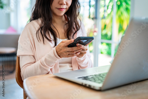 Asian female freelancer holding a mobile phone, messaging or checking notifications in front of a laptop