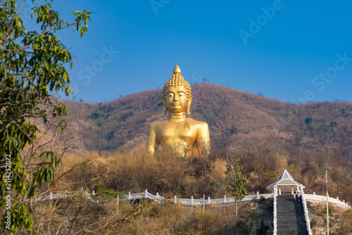 Golden Buddha Statue on Hillside: A majestic, giant golden Buddha statue sits serenely atop a lush green hillside, overlooking a landscape of trees and mountains under a clear blue sky.