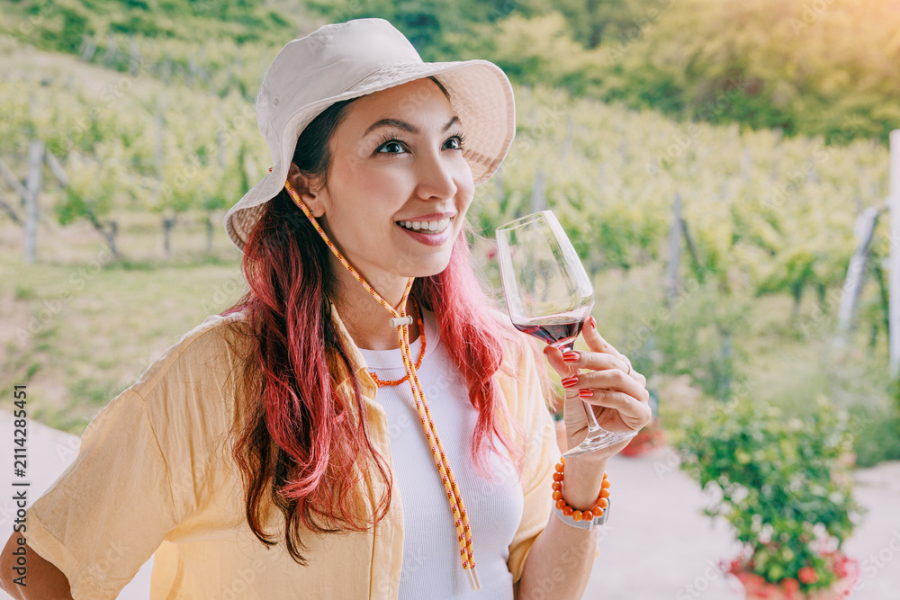 Obraz premium Happy young woman holding red wine glass at a sunny vineyard. Degustation tourism concept.