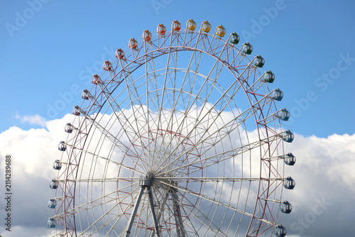 Colorful Ferris wheel gondolas against bright blue sky and clouds.