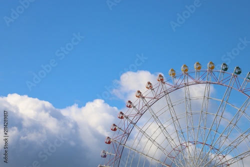 Colorful Ferris wheel gondolas against bright blue sky and clouds.
