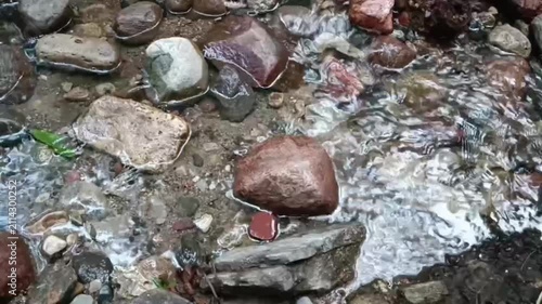 Clear shallow stream water flows gently over smooth wet rocks and pebbles of various sizes and colors. Small ripples and reflections glisten on the water surface