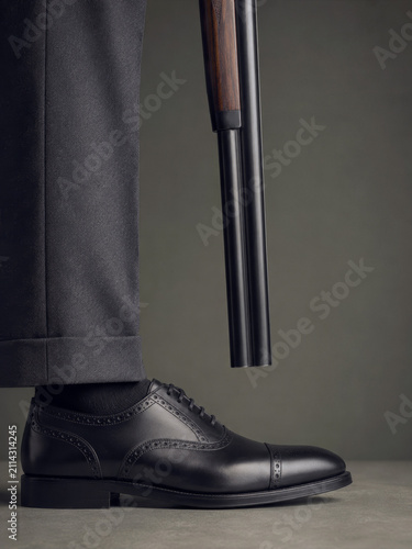 Humorous metaphor of undermining oneself with business man in black formal trousers and leather oxford shoe holding a barrel toward his foot