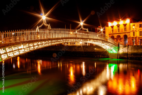 Canvas Print Night view of the Ha'penny Bridge in Dublin, its ironwork illuminated in gold and green, reflected in the River Liffey, with historic buildings glowing warmly in the background