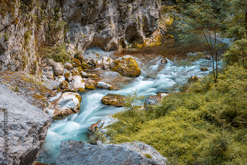 Fotografie Clear river water swiftly flowing through a narrow mountain gorge, surrounded by