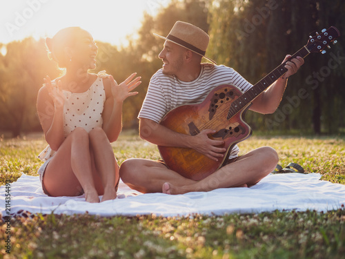 A young couple embraces the magic of golden hour with an outdoor jam session in a scenic park. Sitting comfortably on a picnic blanket, they enjoy live music, laughter, and each other's company.