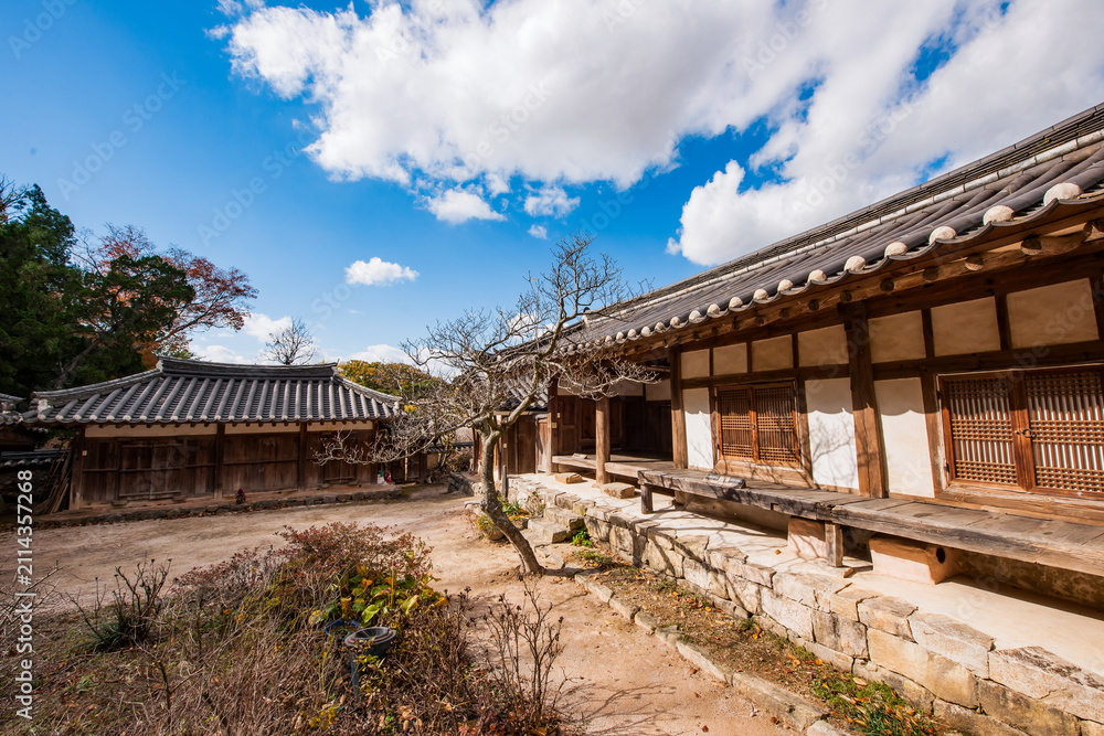 Obraz premium Traditional Korean Hanok House Featuring Wooden Structure, Stone Foundation, and Tiled Roof Set Against a Dynamic Blue Sky and White Clouds