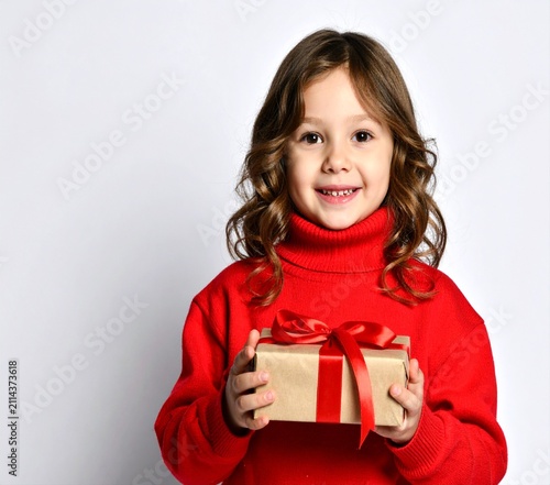 Smiling young girl in a red knitted turtleneck sweater holding a wrapped Christmas gift box with ribbon.