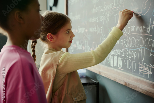 Foto Caucasian schoolgirl writing code on classroom chalkboard while Black classmate observing, showing early STEM learning and teamwork