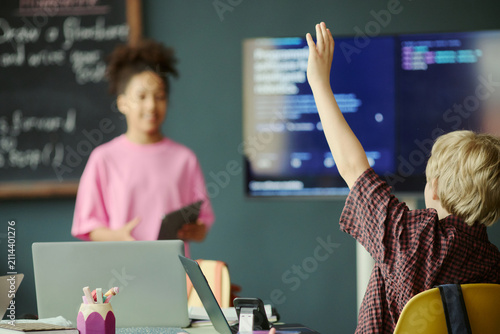 Принти на полотні Schoolboy raising hand during coding lesson while girl classmate presenting near digital screen, showing classroom engagement