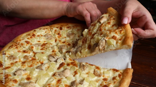 A woman's hands grasp a delicious triangular slice of round Hawaiian pizza with a fluffy edge. The filling consists of juicy chicken breast, diced pineapple, and baked, melted mozzarella cheese.