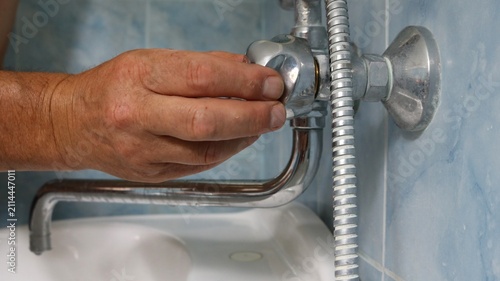 A man's hand adjusts or repairs a chrome faucet valve in a bathroom. The long spout and the edge of a white sink are visible below, and a shower hose is visible to the right. 