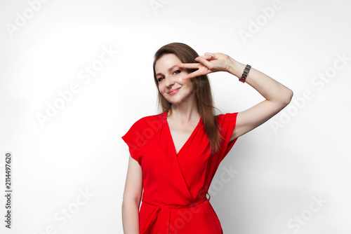 Young girl in red dress showing peace gesture near eye. Isolated on white background.
